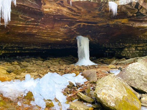 A cave interior with icicles hanging from the ceiling and a large ice formation on the ground among rocks.