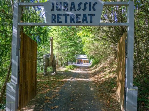 A sign reading "Jurassic Retreat" at the entrance of a wooded path, with a dinosaur statue nearby.