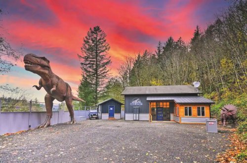 A dinosaur statue stands near a building surrounded by trees, under a vibrant sunset sky.