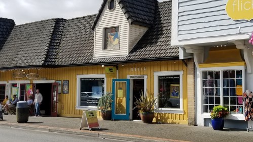 A colorful storefront with a yellow facade, large windows, and a blue door, featuring plants and people outside.