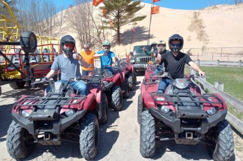 A group of people on ATVs, smiling and posing for the camera, with a sandy dune in the background.