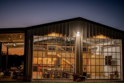 A modern building with large windows, showcasing a warm interior and a staircase, illuminated at dusk.
