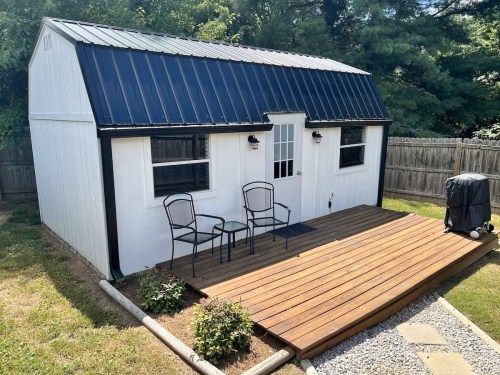 A small white shed with a black metal roof, featuring a wooden deck and two chairs in a grassy yard.