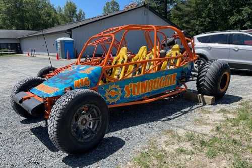 A colorful dune buggy with an orange frame and yellow seats parked on gravel, with a building and portable restroom in the background.