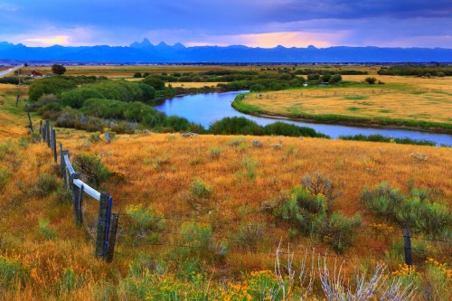 A serene landscape featuring a winding river, grassy fields, and distant mountains under a colorful sky.
