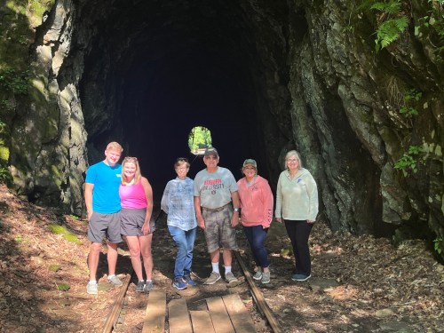A group of six people stands at the entrance of a rocky tunnel, surrounded by greenery and a dirt path.