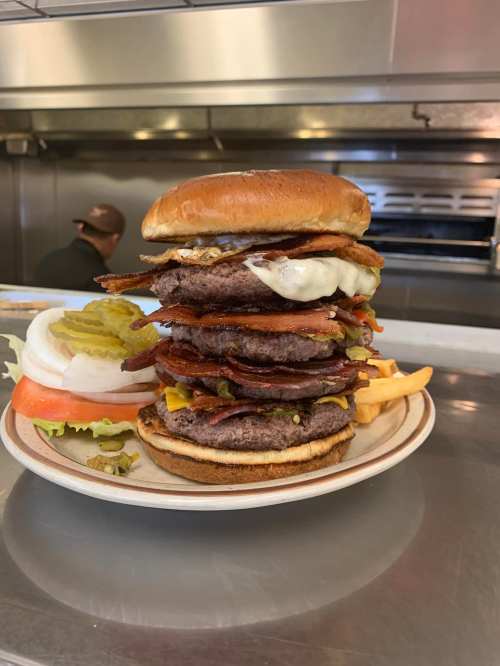 A towering burger stacked with multiple patties, bacon, cheese, lettuce, tomato, pickles, and a toasted bun.