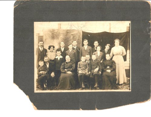 A vintage black-and-white group photo of 18 people, including men, women, and children, posed together indoors.