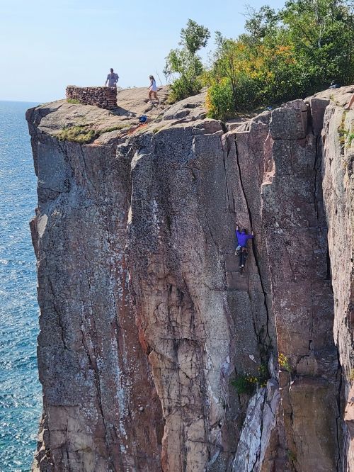 A climber ascends a rocky cliff by the ocean, while two people relax on top of the cliff in the background.