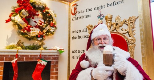 Santa Claus in a festive setting, holding a drink with whipped cream, surrounded by Christmas decorations.