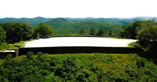 Aerial view of a large, empty stone platform surrounded by lush green hills and trees under a bright sky.