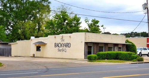 A single-story building with "The Backyard Bar-B-Q" sign, surrounded by greenery and parked cars on a street.