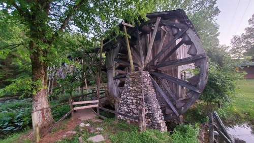 A rustic water wheel beside a stone structure, surrounded by lush greenery and a serene landscape.