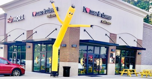 A yellow inflatable tube man waves outside a restaurant with a playful sign, featuring a red heart and cheese graphics.