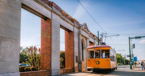 A vintage yellow streetcar passes through a historic archway on a sunny day.