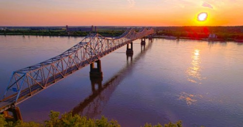 A bridge spans a river at sunset, with vibrant colors reflecting on the water and a clear sky in the background.