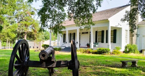 A historic white house with green shutters, featuring a cannon in the foreground and surrounded by trees and grass.