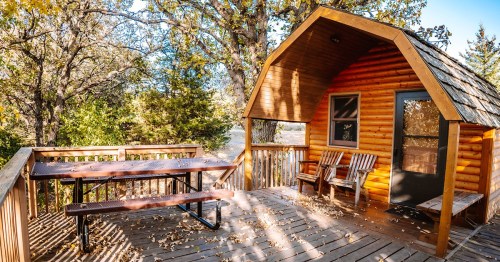 Cozy wooden cabin with a deck, surrounded by trees, featuring picnic tables and chairs.