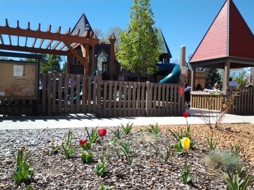 A playground with a wooden structure, slide, and colorful tulips in the foreground under a clear blue sky.