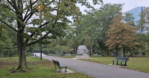 A park scene featuring benches, trees, and a large sculpture of a head in the background on a cloudy day.