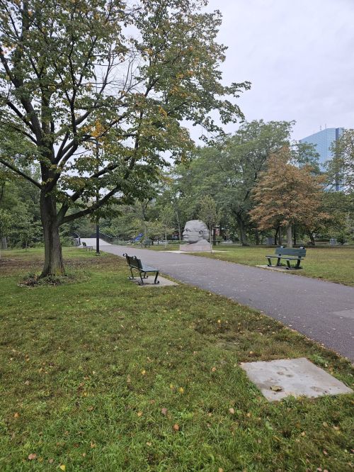 A park path lined with benches, trees, and a large sculpture in the distance under a cloudy sky.