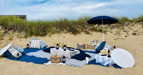 A cozy beach picnic setup with blankets, pillows, umbrellas, and baskets on sandy dunes under a blue sky.