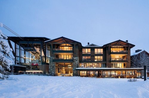 A modern lodge with large windows, surrounded by snow, illuminated at dusk.