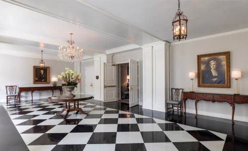 Elegant foyer with black and white checkered floor, chandeliers, and classic furniture, featuring a portrait on the wall.