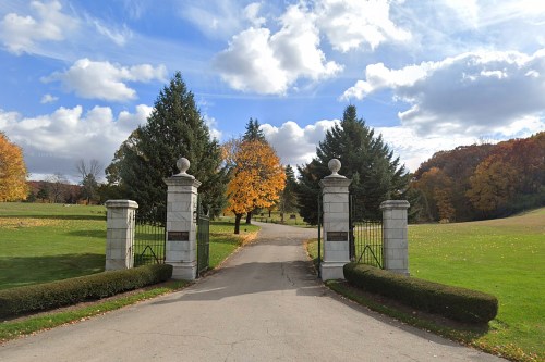 A scenic entrance with stone pillars and gates, leading into a lush green park with autumn trees under a cloudy sky.