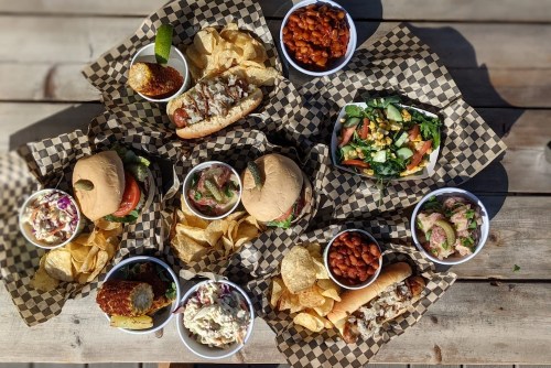 A spread of various sandwiches, sides, and salads on checkered paper, served with chips on a wooden table.