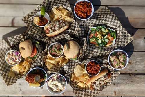 A spread of various sandwiches, sides, and salads on checkered paper, served with chips on a wooden table.