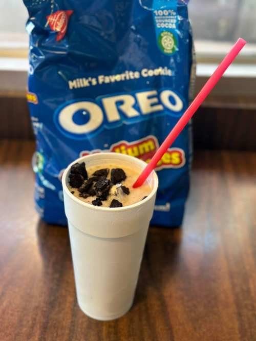 A white cup filled with a creamy Oreo milkshake, topped with crushed Oreos, in front of a bag of Oreo cookies.