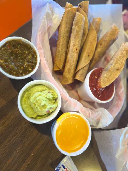 A basket of crispy taquitos with bowls of guacamole, salsa, and cheese sauce on a wooden table.