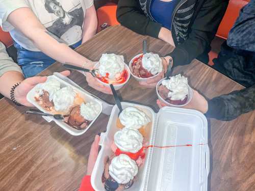A group of hands holding dessert cups topped with whipped cream and syrup on a wooden table.