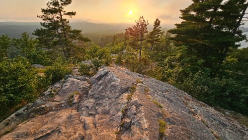 Sunset over a rocky landscape, surrounded by trees and distant mountains, creating a serene natural scene.