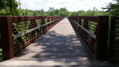 A wooden bridge stretches into a green landscape, surrounded by trees under a partly cloudy sky.