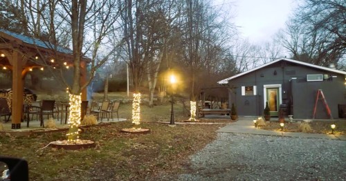 A cozy outdoor space with string lights, a small building, and a gravel path surrounded by trees at dusk.