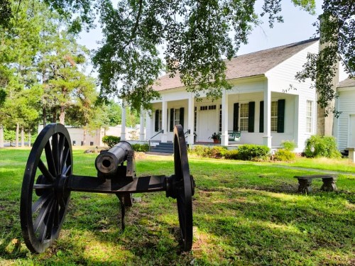 A historic white house with green shutters, featuring a cannon in the foreground and trees providing shade.