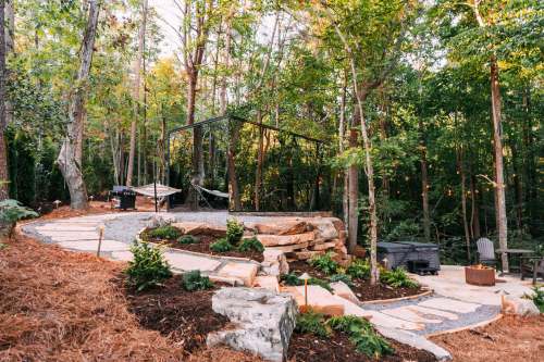 A serene outdoor space with a stone path, hammock, and seating area surrounded by trees and greenery.