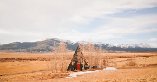 A-frame cabin with a bright red door in the Colorado Mountains with the Front Range visible in the background, an open field, and sparse trees.