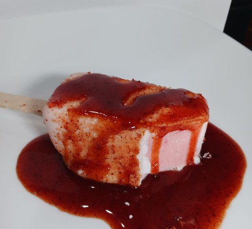 A pink popsicle on a plate, drizzled with a red sauce, resting on a white background.