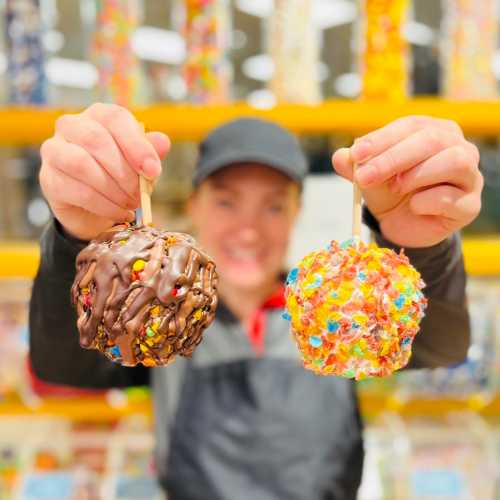 A person holds two colorful, decorated candy apples, one covered in chocolate and sprinkles, the other in bright candies.