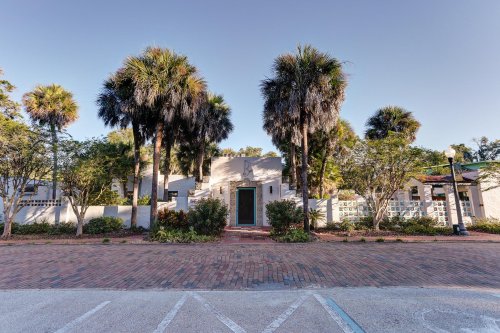 A modern building surrounded by palm trees and greenery, with a brick pathway leading to the entrance.