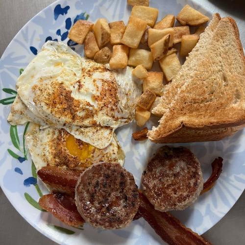 A plate with two fried eggs, crispy bacon, sausage patties, toast, and golden-brown potato cubes.