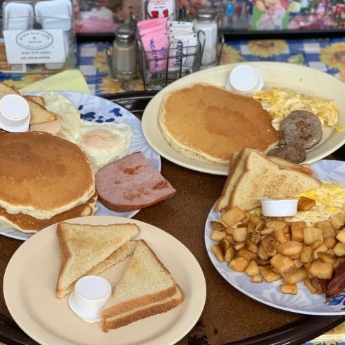 Two plates of breakfast featuring pancakes, eggs, sausage, toast, and home fries, served with condiments.
