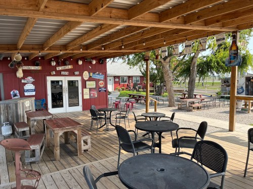 Outdoor seating area of a rustic bar with wooden tables, chairs, and a red building in the background.