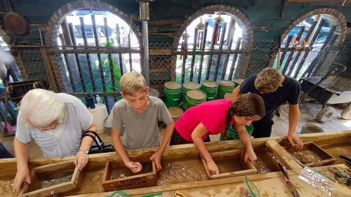 Four people, including an older woman and three children, sift through water in wooden boxes, searching for gems or minerals.