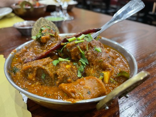 A bowl of rich, spicy curry with meat, garnished with herbs and red chilies, served on a wooden table.