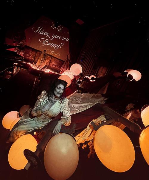 A ghostly figure in a vintage dress sits among glowing orbs, with a sign asking, "Have you seen Bunny?" in a dimly lit room.