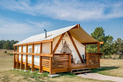 A spacious glamping tent with wooden deck and railings, set in a grassy area surrounded by trees under a blue sky.